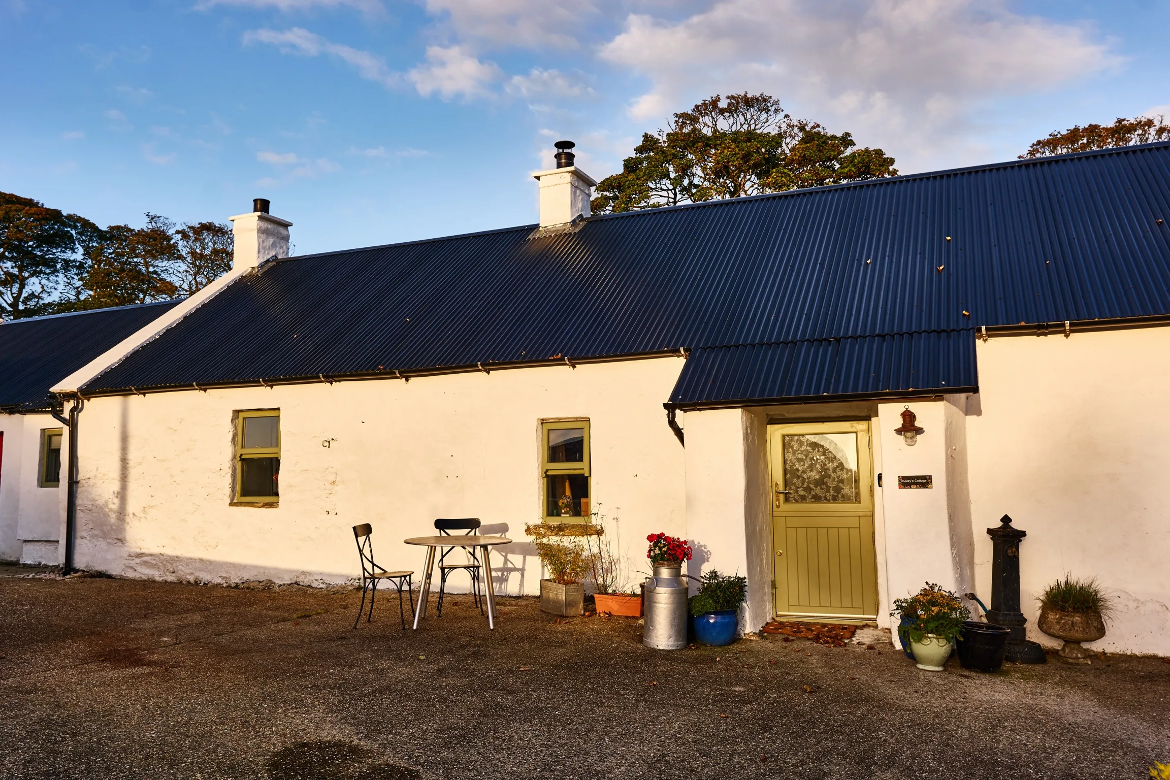 The whitewashed cottage with its dark corrugated roof and green half-door in golden evening light, a bistro table and a milk churn of red geraniums out front