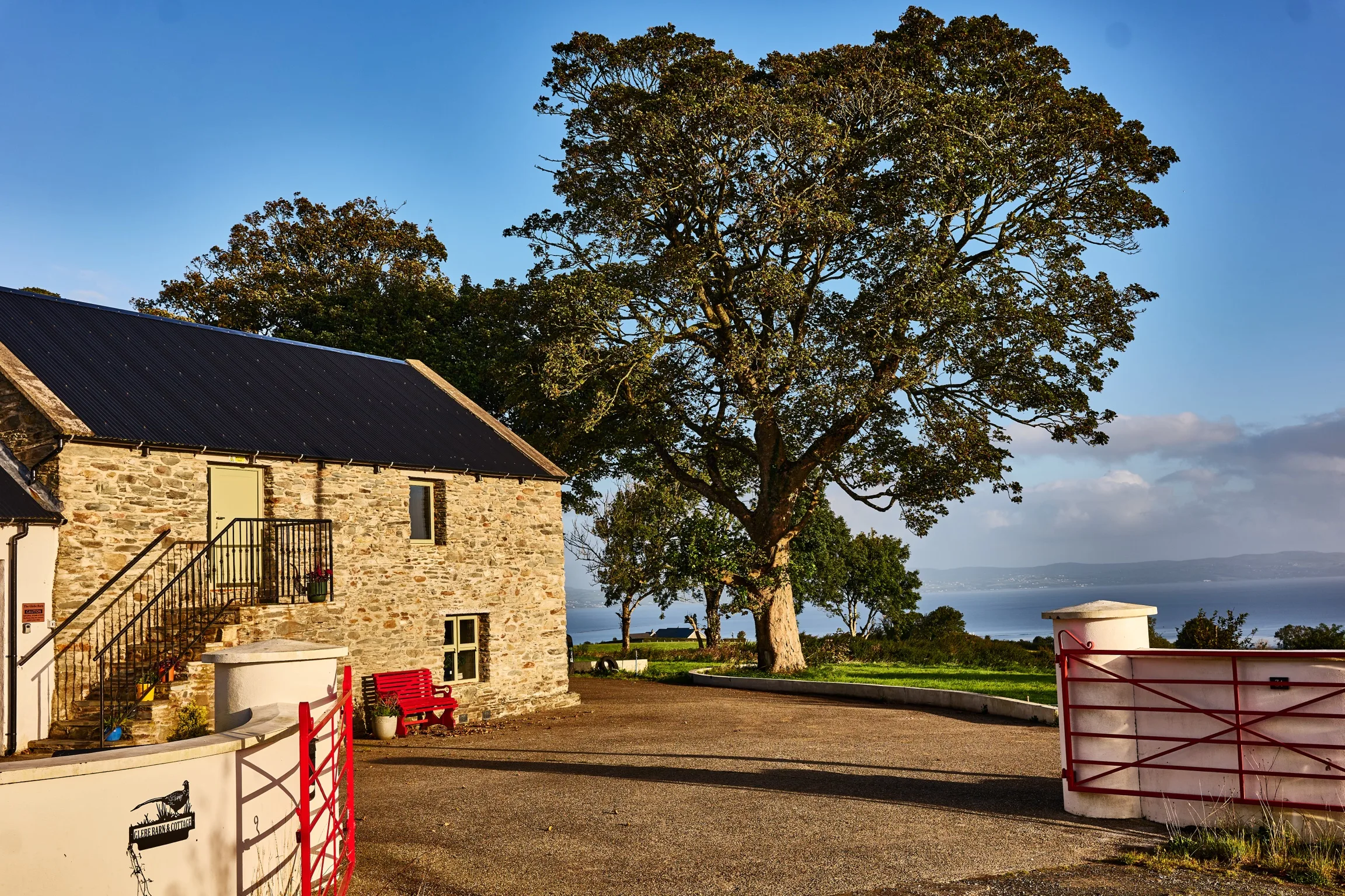 The red Glebe Barn and Cottage gate with its pheasant sign, the stone barn and the great oak behind, Lough Foyle in the distance
